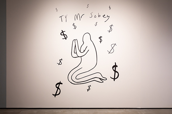 A photo of an art installation, depicting a person on their knees with their hands pressed together. Above their head is the words "TY MR SOBEY" and dollar signs.