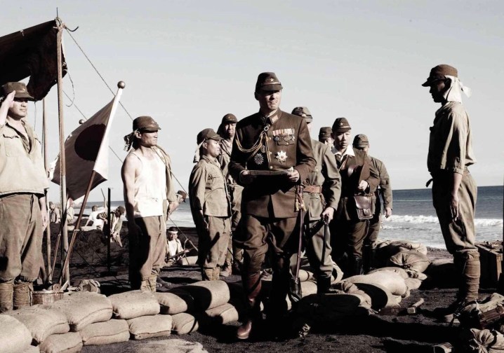 A group of Japaneze soldiers stand on a beach as one walks into a bunker.