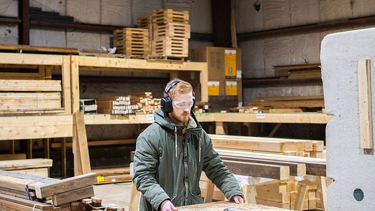 A photo of a man in a hoodie, wearing ear and eye protection and feeding wood through a machine