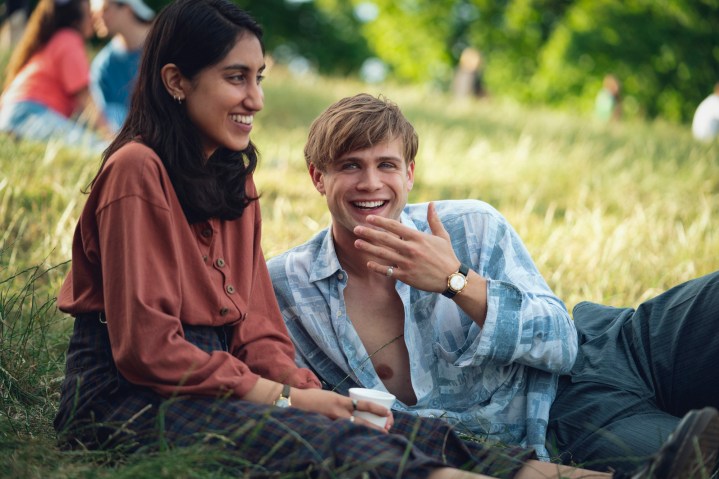 A boy lays down next to a girl siting in a field.