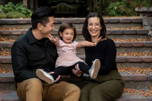 A couple wearing dark shirts, sitting on outdoor stone steps and holding a happy little girl in a pink dress and sneakers