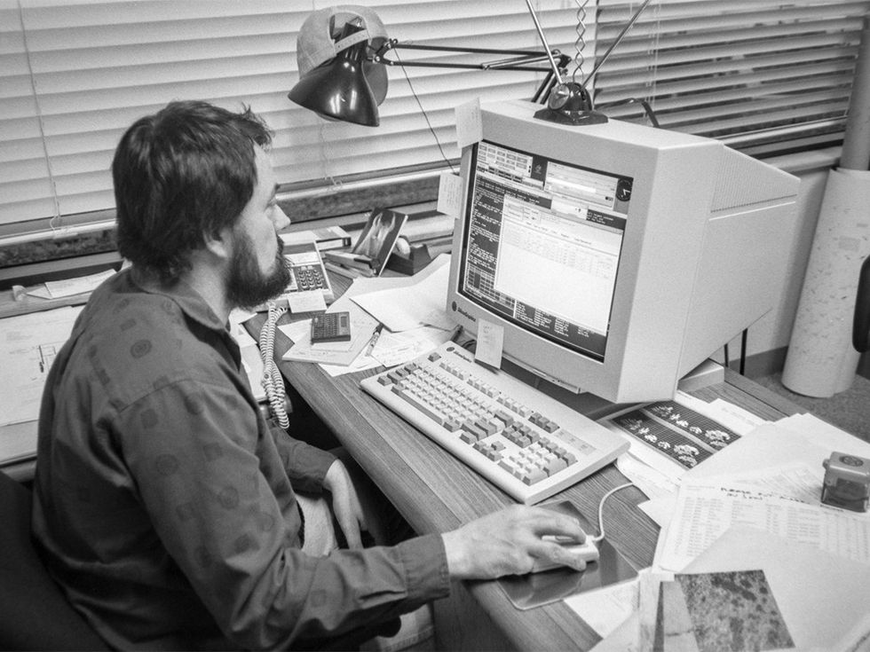 A black and white photo of a bearded man sitting in front of an old computer.