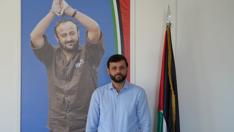 A bearded man in a blue shirt stands in front of a portrait of a man in handcuffs with his arms raised. 