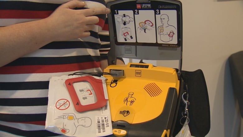 Someone's hand holding open an AED kit, showing its contents: pads and images showing where to place the pads. 