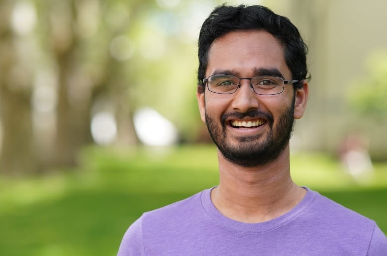 Man with black hair and beard wearing a purple shirt on a blurred outdoor background.