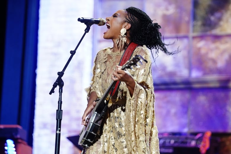 Allison Russell, a Black woman with long, dark, braided hair, sings enthusiastically into the microphone while playing her guitar.