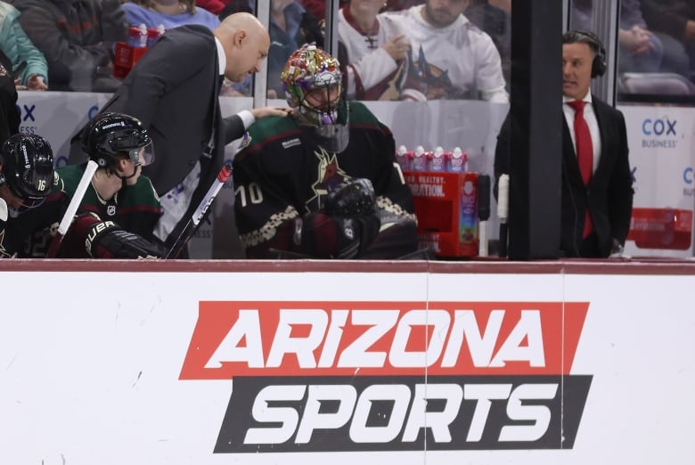 A hockey coach in a suit speaks with a goaltender, while placing his left hand on the goalie's right shoulder.