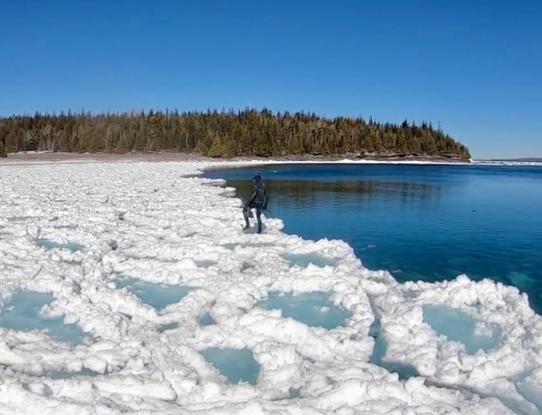 A man in a wetsuit walks on an icy shore of a blue lake. 