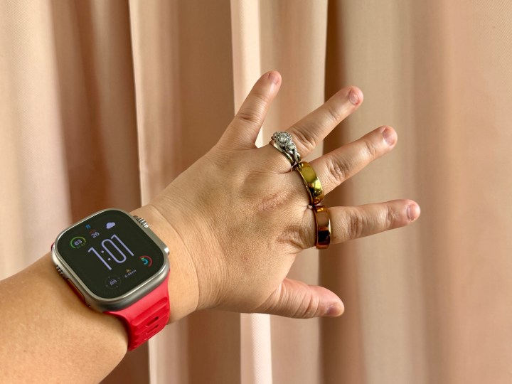 An Apple Watch Ultra with red strap on wrist with a Movano Evie and Oura Ring on fingers.