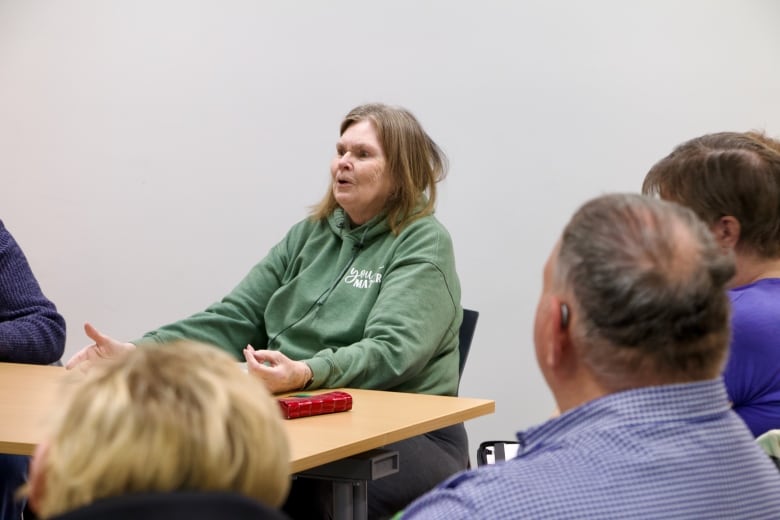 An older woman sits at a table, talking to a circle of people.