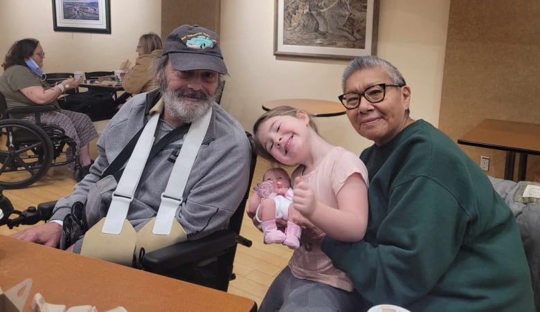 A man with a beard sits in a wheelchair with his arm in a sling, with a woman with short hair sitting next to them, holding a smiling child between them.