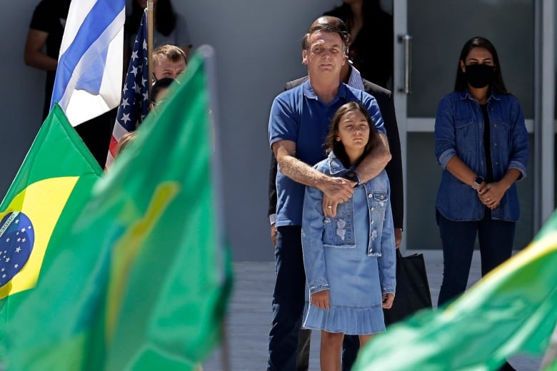 Jair Bolsonaro stands with his arms wrapped around the shoulders of his young daughter, surrounded by green, yellow and blue Brazilian flags. 