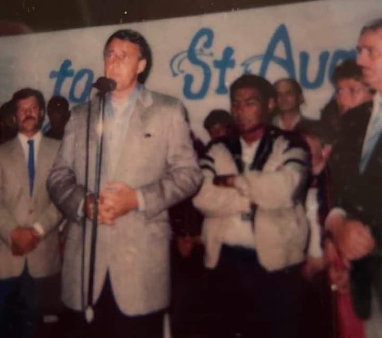 A group of people stand behind a man speaking at a microphone