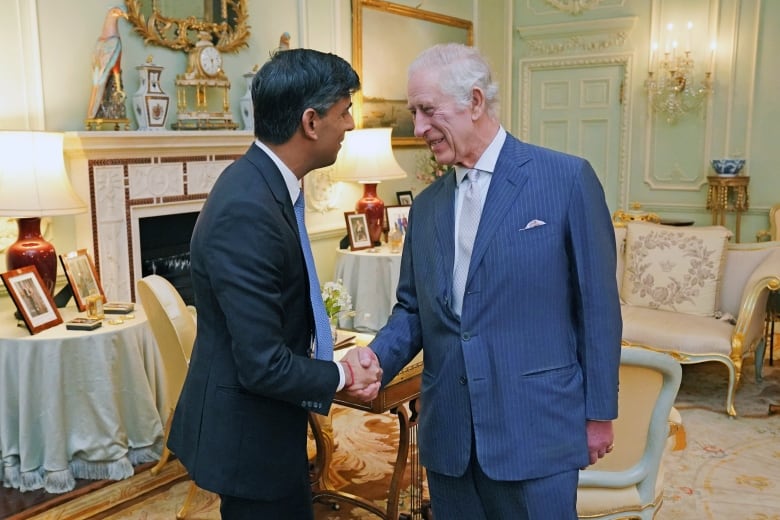 Two people shake hands while standing in a sitting room with upholstered furniture.