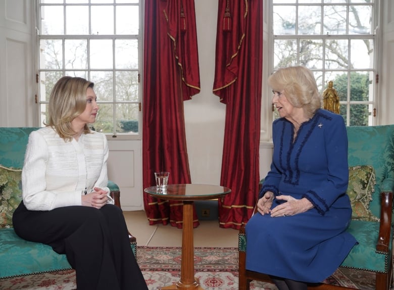 Two women sit in parlour chairs, talking to each other.