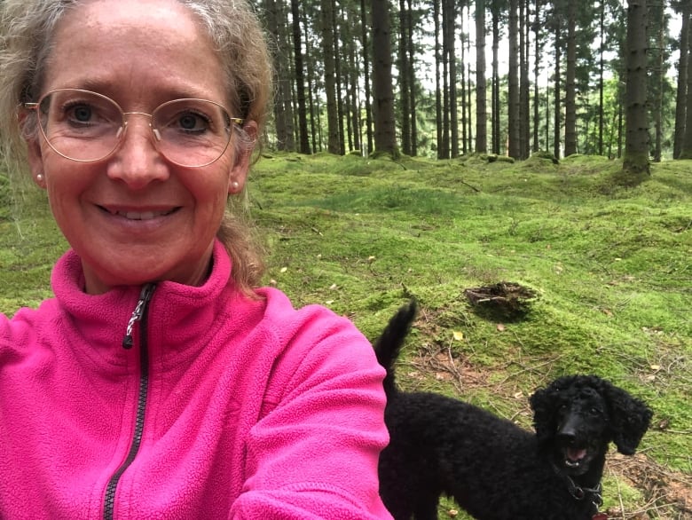 A woman in a pink fleece sweater poses for a selfie with a black dog. A mossy ground and trees are seen in the distance.