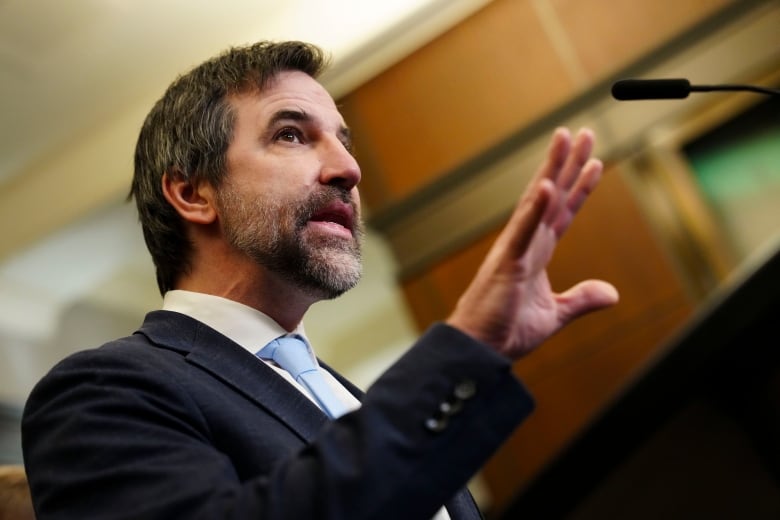 A man wearing a suit with a beard talks at a podium.