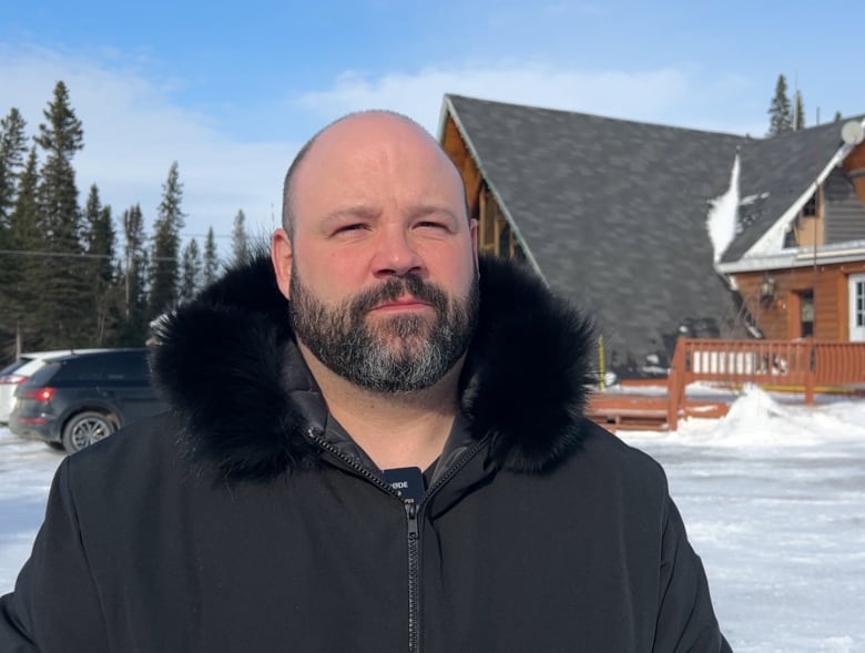 A bald man with a beard stands in front of a ski lodge.