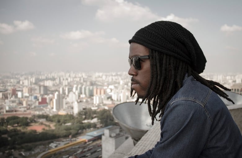 Chris Strikes looks out over a city view. Strikes' hair is styled in locs that are slightly past his shoulders.