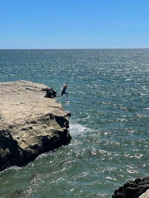 Photo taken from a distance of a woman leaping off a cliff into the ocean on a sunny day.