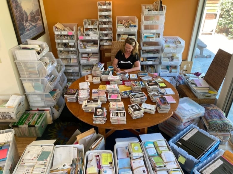 A woman sits in an office at a table writing. The table, and the entire room around her, are stacked with boxes upon boxes of letters.