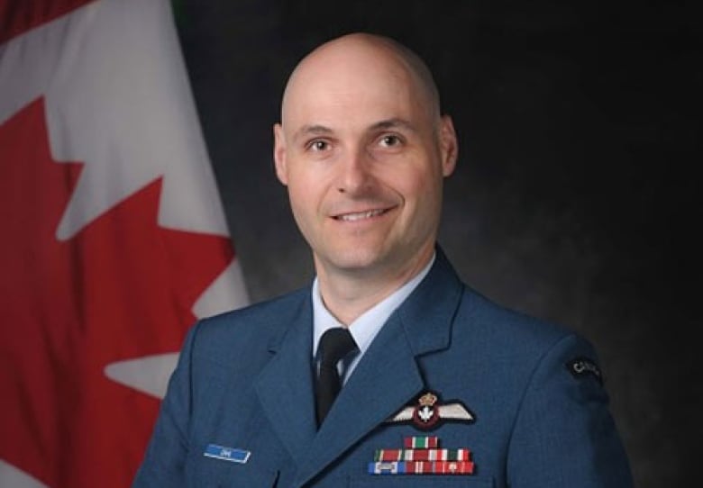 A bald man in a blue military uniform smiles in a posed photo in front of a Canadian flag.