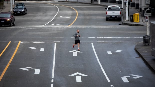 A jogger in a grey hoodie, black shorts and running shoes crosses an otherwise deserted three-lane street in Vancouver.