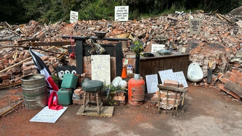 A display of broken chairs, pieced of a bar, plastic containers and a urinal is erected in front of a pile of debris. Throughout the debris are handwritten signs that read, "Rise from the ashes," More than a pub," and "A crime against the Black Country — happy hour is over."