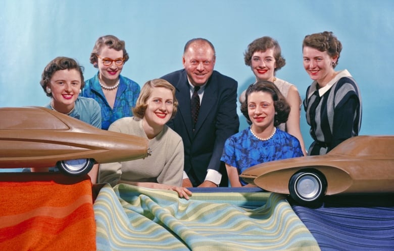 Six women and one man smile for a posed photo with vehicle models.