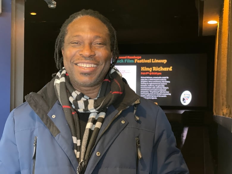 Black man with dreadlocks wearing a winter coat stands in a theatre lobby