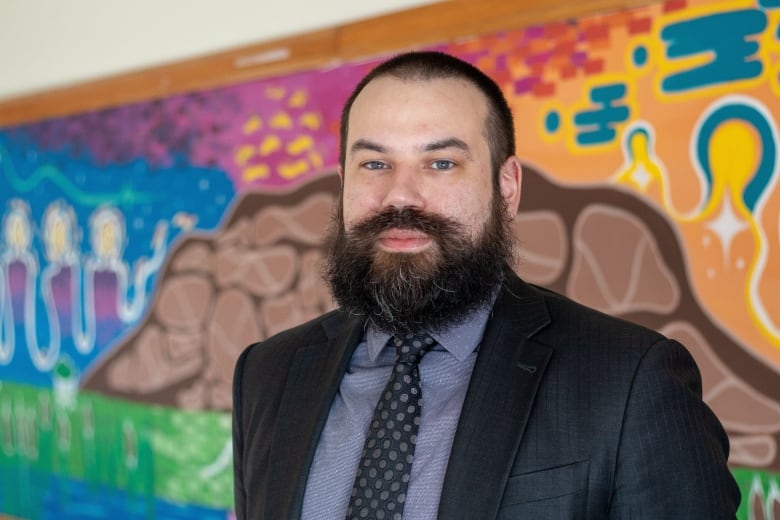 A person wearing a suit and grey shirt stands in front of a multi-colour mural.