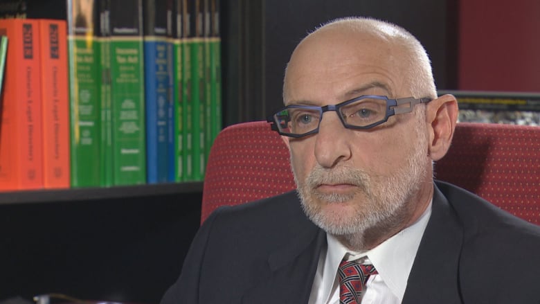 A bald man with blue geometric eyeglasses is pictured in front of a bookcase, wearing a suit.