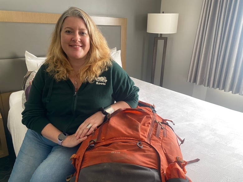 A woman in a green Roots hoodie is sitting on a bed and leaning on a orange hiking backpack. 