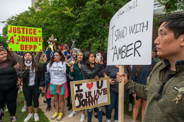 A man on the right side of the frame holds up a sign reading "I stand with Amber" as a crowd of people behind him cheer and hold signs in support of actor Johnny Depp.