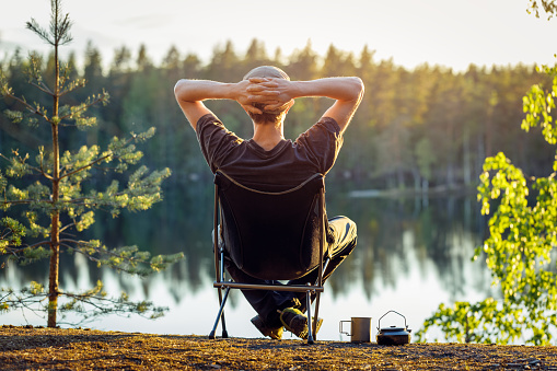 Der Mann sitzt an einem schönen Sommerabend in einem Campingstuhl auf dem Hintergrund eines Waldsees.