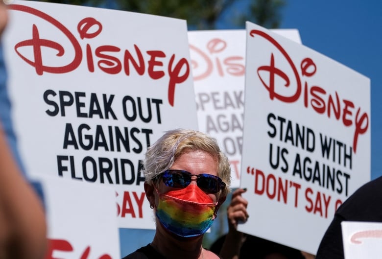 A person in sunglasses and a mask stands in front of picket signs that say Disney, stand with us against Don't Say Gay.