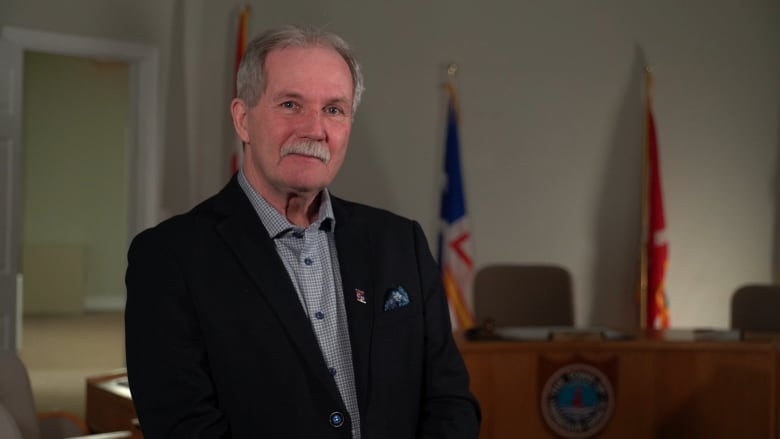 Portrait of a man with a grey moustache standing in a room with the Newfoundland and Labrador flag in the background.