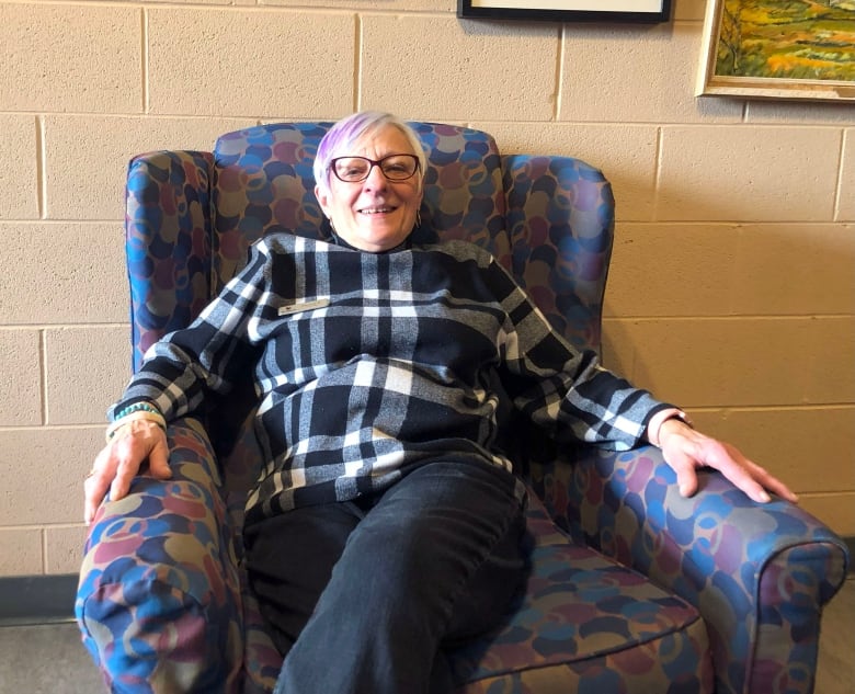 A woman with white hair dyed with a purple streak poses on a patterned armchair.