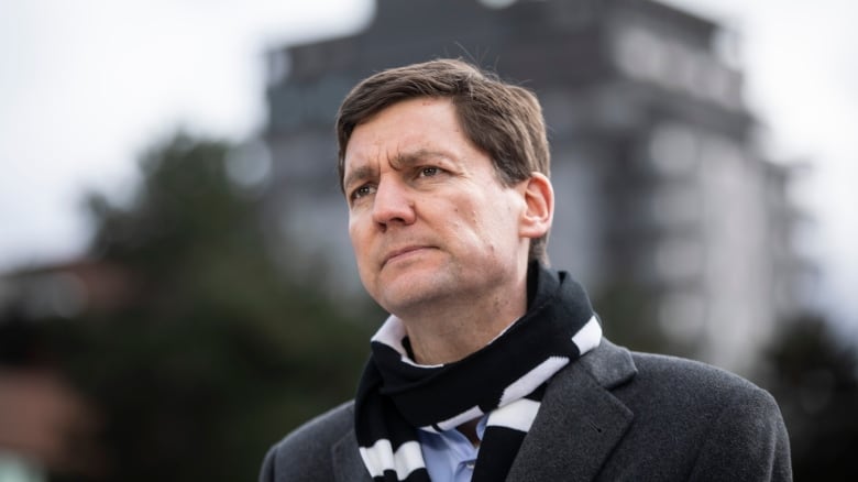 A tall white man wearing a black scarf looks on, with a large building in the background.