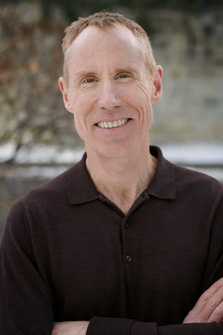 Manns is wearing a long sleeved brown collared shirt. He has his arms crossed and is smiling as he looks into the camera. He's standing outside.