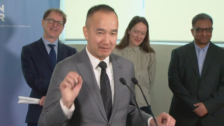 A person in a suit and tie speaks at a podium with politicians behind him and signs reading "B.C. Cancer."