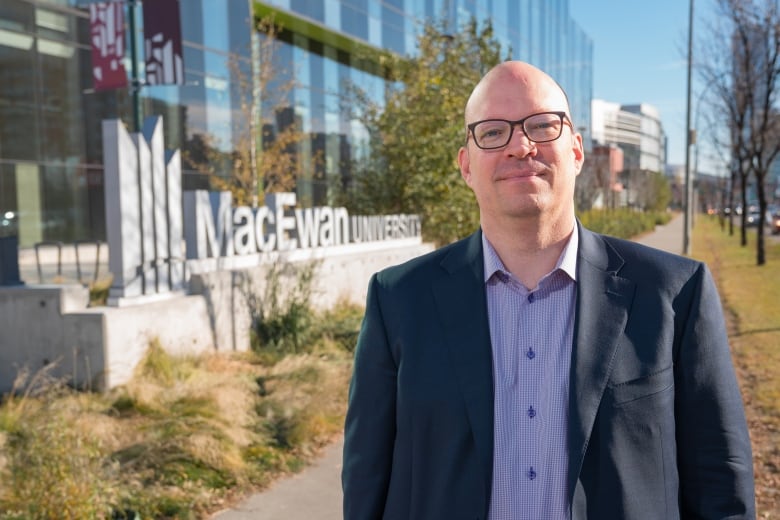 A bald man with glasses stands outside a glass building on a university campus on a sunny day, with a sign reading MacEwan University on top of a concrete platform behind him.