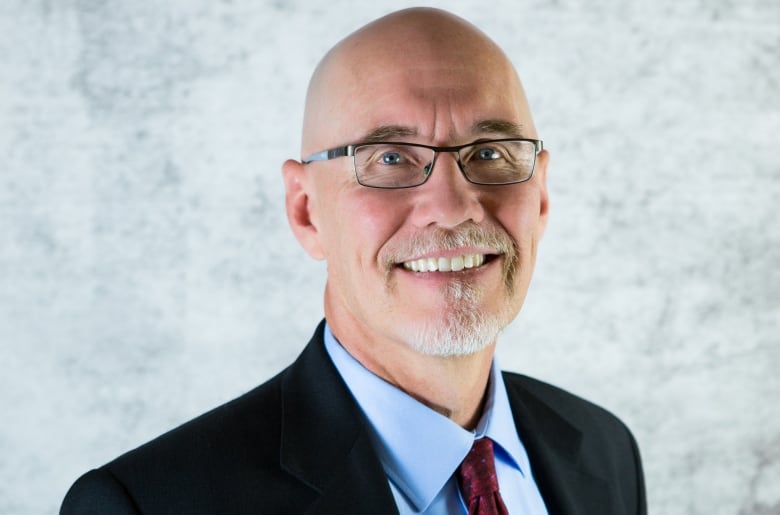 Bald man with glasses, wearing a suit and tie and smiling at the camera. 