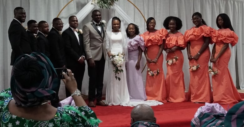 A newly married couple stands under a lace and flowered arch, surrounded by suit-clad men on one side and women in peach-coloured dresses on the other.