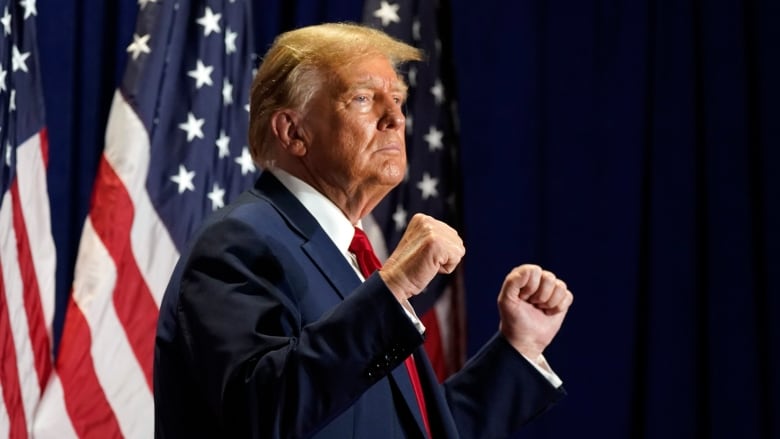 An older, cleanshaven man in a suit and tie stands with fists raised on a stage, with a backdrop of at least three American flags.