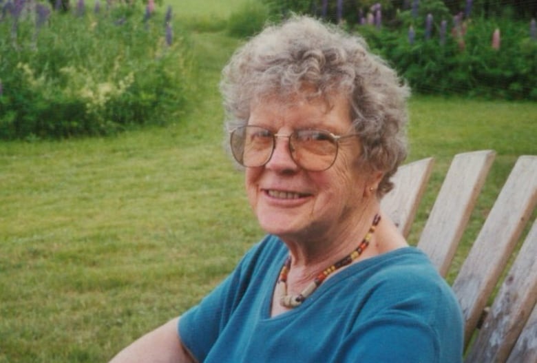 A woman with glasses and short grey hair sits outside in a lawn chair. 