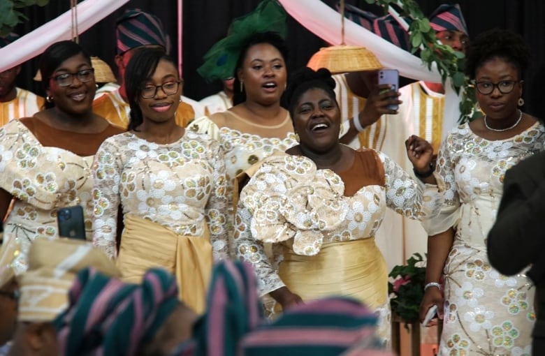 Five smiling women wearing cream and white dresses stand in a line.