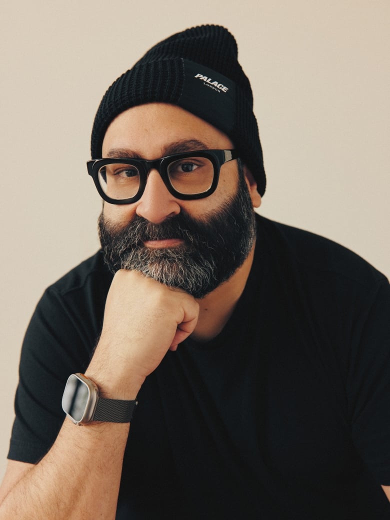 Headshot of a south Asian ban with a tidy greying beard. He is wearing a black tshirt, blacktoque, and glasses with black frames. 