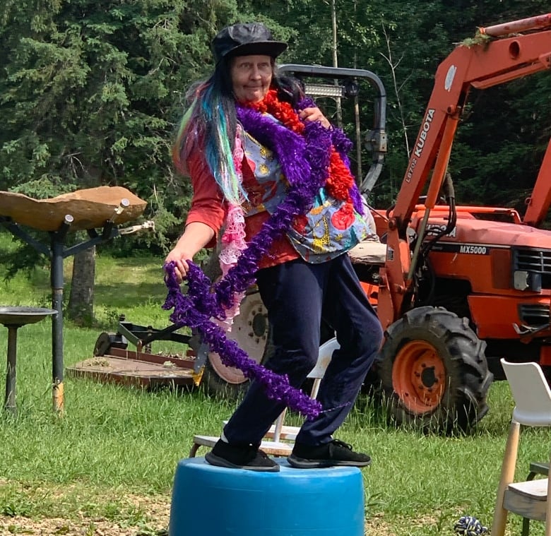 A woman wearing purple and red feather boas and wearing a jaunty hat dances while standing on top of a blue barrel. 