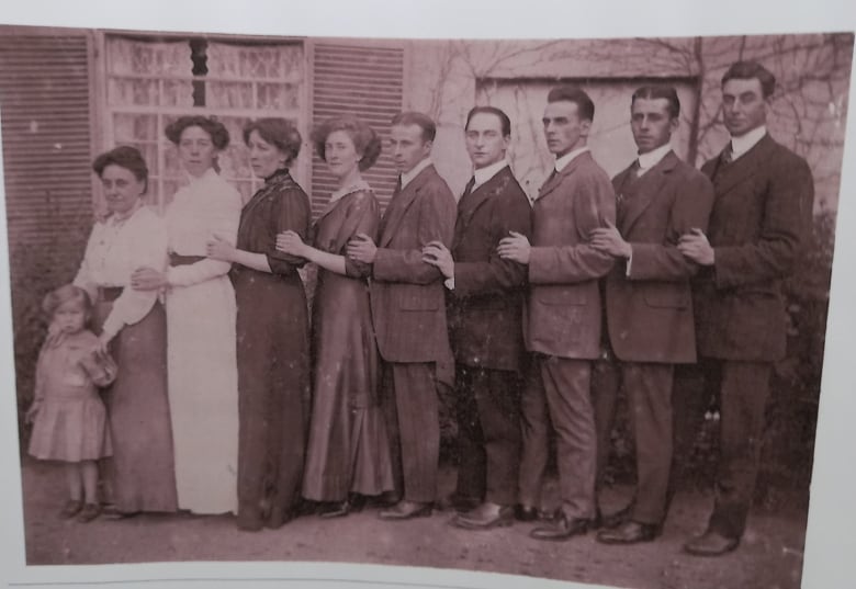 A group of people stand in a line. There is a child on the far left, a few women, and then a few men. The photo is more than 100 years old.